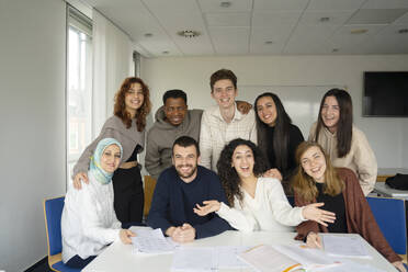 Fröhliche Schüler sitzen um einen Tisch mit Büchern im Klassenzimmer - NJAF00460