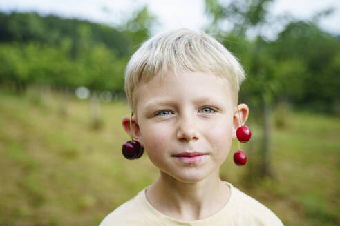Blonder Junge mit Kirschen an den Ohren im Garten - NJAF00466