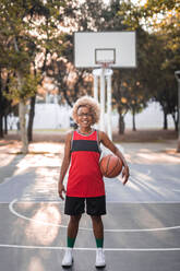 Full body of young African American female basketball player in sportswear with ball standing on sports ground in park and looking at camera - ADSF45742