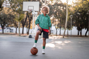 Full body of cheerful African American female basketball player in cozy jacket and shorts with leg over ball while standing in basketball court - ADSF45745