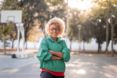 Happy young African American female in sport jacket and glasses crossing arms and looking at camera while standing in park on sunny day - ADSF45746