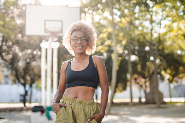 Happy young African American female in glasses standing with hands in pockets on sport ground on sunny day in park while looking at camera - ADSF45753