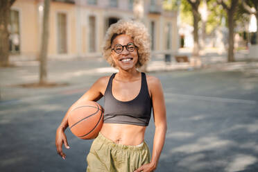 Positive young African American female basketball player in ethnic clothes and glasses holding ball and looking at camera while standing on street - ADSF45754