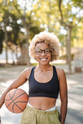 Positive young African American female basketball player in ethnic clothes and glasses holding ball and looking at camera while standing on sports ground - ADSF45756