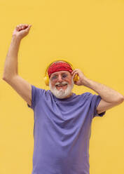 Happy bearded elderly man in grey hair and casual clothes with red scarf and headphones smiling with closed eyes enjoying music and dancing with hand up - ADSF45761