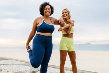 Zwei unterschiedliche junge Frauen in Sportkleidung lächeln, während sie sich auf der Promenade dehnen und aufwärmen. Interrassische Freundinnen genießen ihr Workout am Strand mit den Wellen des Meeres im Hintergrund. - JLPSF30597