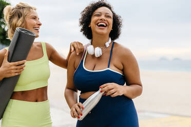 Junge, fitte Frauen in Sportkleidung lachen und lächeln, während sie auf einer Promenade in der Nähe des Strandes stehen. Fitness-Freunde, die nach einem gesunden Workout gemeinsam ihre Yoga-Utensilien in der Hand halten. - JLPSF30601