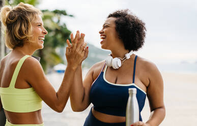 Zwei fitte Frauen in Sportkleidung stehen an der Strandpromenade und klatschen sich gegenseitig ab, um ihren gesunden und aktiven Lebensstil zu feiern. Fitness-Frauen, die ein Trainingsprogramm im Freien genießen. - JLPSF30604