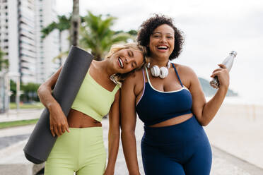 Porträt von zwei Fitness-Freundinnen, die mit ihren Yoga-Geräten in der Hand am Meer stehen und in die Kamera lächeln. Glückliche junge Frauen mit unterschiedlichen Körpergrößen, die einen aktiven Lebensstil pflegen. - JLPSF30605