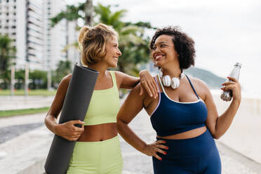 Durchtrainierte Frauen in Sportkleidung lächeln und lachen gemeinsam mit einer Yogamatte und einer Wasserflasche in der Hand auf einer Strandpromenade. Sie genießen die Gesellschaft des anderen, um sich im Freien zu bewegen und einen gesunden Lebensstil zu pflegen. - JLPSF30606
