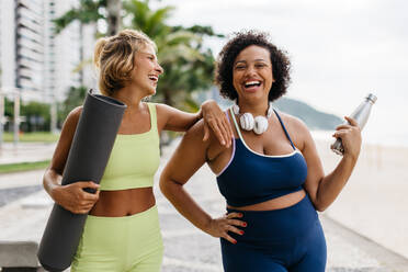 Zwei Frauen in Sportkleidung stehen auf einer Strandpromenade und halten eine Yogamatte und eine Wasserflasche in der Hand. Glückliche Yoga-Freundinnen lachen zusammen am Meer und genießen gemeinsam einen gesunden Lebensstil. - JLPSF30607