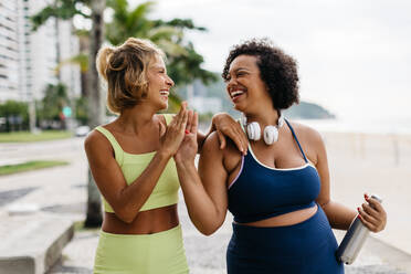 Sportliche Frauen in Fitnesskleidung, die sich gegenseitig abklatschen, um eine erfolgreiche Trainingseinheit im Freien zu feiern. Zwei Fitness-Freundinnen mit unterschiedlichen Körpergrößen, die gemeinsam einen gesunden Lebensstil pflegen. - JLPSF30608