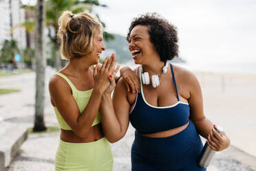 Zwei glückliche Frauen in Sportkleidung genießen ihr Trainingsprogramm an der Strandpromenade und feiern es mit einem High-Five. Freundinnen mit unterschiedlichen Körperformen, die einen Lebensstil der Fitness pflegen. - JLPSF30609