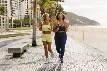 Zwei Läuferinnen joggen entlang der Strandpromenade am Meer und genießen ihr Trainingsprogramm. Fitness-Frauen lächeln und plaudern fröhlich, während sie Ausdauertraining machen, beide tragen Sportkleidung für ihren aktiven Lebensstil. - JLPSF30610