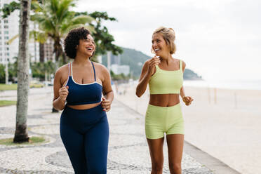 Two happy young women in fitness clothing jog together on the beach promenade. Female runners with diverse body shape laugh together by the ocean, enjoying a perfect summer exercise routine. - JLPSF30611