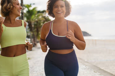 Fröhliche junge Frauen joggen in Fitnesskleidung an der sonnigen Strandpromenade entlang. Zwei Läuferinnen, die die Vielfalt der Körper repräsentieren, genießen ein gesundes Trainingsprogramm im Freien. - JLPSF30612