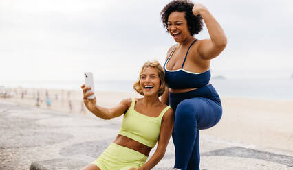 Zwei Frauen mit unterschiedlichen Körpertypen feiern ihren Trainingserfolg, indem sie ein Selfie an der Strandpromenade machen - JLPSF30613
