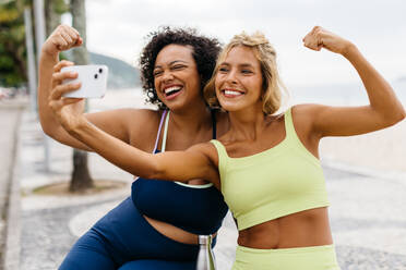 Two women in sportswear take a selfie with a smartphone, smiling and showing off their healthy biceps. Friends celebrating a healthy lifestyle after a great workout on the ocean promenade. - JLPSF30621