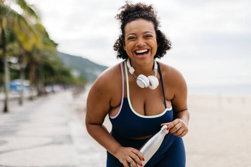 Glückliche junge Frau, die auf einer Strandpromenade in der Nähe des Ozeans steht, eine Sportwasserflasche in der Hand hält und aufgeregt lacht. Gesunde junge Frau, die in Fitnesskleidung ein Strandtraining absolviert. - JLPSF30628
