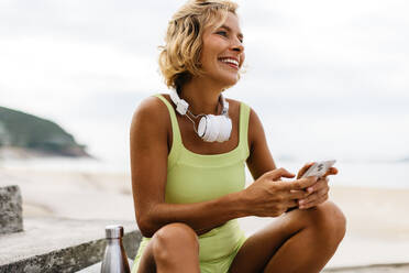 Kaukasische Frau in Trainingskleidung macht eine Pause von ihrer aktiven Routine im Freien und benutzt ihr Telefon, um im Internet zu surfen. Glückliche junge Frau, die Technologie benutzt, um ihre Fitnessreise zu navigieren. - JLPSF30632