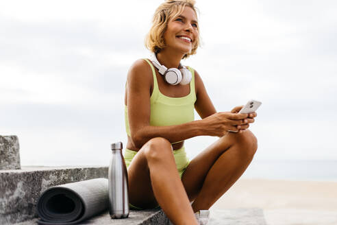Eine Frau surft in einer Fitness-App auf ihrem Handy, genießt die Meeresbrise und sucht nach Inspiration für ihre bevorstehende Yoga-Praxis. Glückliche kaukasische Frau sitzt mit ihrer Yogamatte auf einer Treppe am Strand. - JLPSF30633