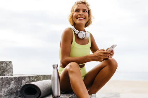 Eine glückliche Frau in Yogakleidung benutzt ihr Smartphone nach einem großartigen Yoga-Workout auf der Promenadentreppe und genießt die Ruhe am Meer und die Erfrischung durch ihr Trainingsprogramm. - JLPSF30634