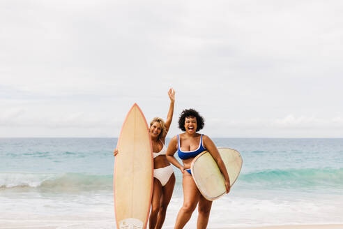 Zwei Freundinnen feiern ein Sommerabenteuer am Strand: Fröhliche Frauen in Badekleidung jubeln aufgeregt, während sie ihre Surfbretter am Ufer halten und gemeinsam auf den Wellen reiten. - JLPSF30641