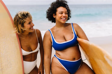 Das Surferleben wird lebendig, wenn zwei Frauen mit ihren Surfbrettern in der Hand fröhlich lächelnd zusammen am Strand stehen. Surferinnen mit unterschiedlichen Körpergrößen genießen ein lustiges Sommererlebnis am Strand. - JLPSF30642