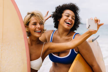 Happy and excited female surfers capture the moment with a selfie on the beach, celebrating a fun holiday and a great surfing destination. Two young women enjoying beach life and beach activities in summer. - JLPSF30643