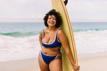 Plus-Size-Frau im Bikini hält ein Surfbrett am Strand, bereit für ein aufregendes Surf-Abenteuer. Weibliche Surferin lächelt in die Kamera und spiegelt die Aufregung des Wassersports und die Freude eines Strandurlaubs wider. - JLPSF30645