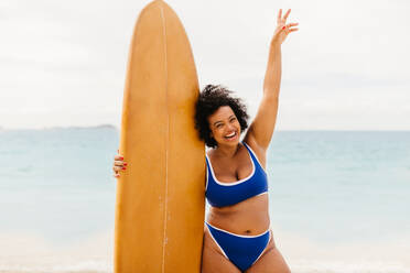 Kurvige junge Frau hält ihr Surfbrett und hebt ihren Arm zur Feier nach einem aufregenden Surfabenteuer an einem sonnigen Strand. Glückliche Plus-Size-Frau erkundet Strandaktivitäten im Bikini. - JLPSF30649