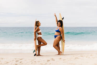 Female surfers in bikinis celebrate after an exciting surfing trip on the beach. Two women with diverse bodies stand with their surfboards on shore, enjoying the surfing life during a summer holiday. - JLPSF30652