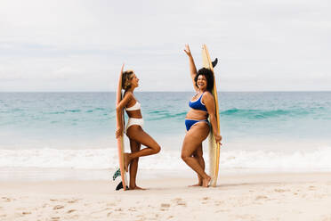Surferinnen in Bikinis feiern nach einem aufregenden Surftrip am Strand. Zwei Frauen mit unterschiedlichen Körpern stehen mit ihren Surfbrettern am Ufer und genießen das Surferleben während eines Sommerurlaubs. - JLPSF30652