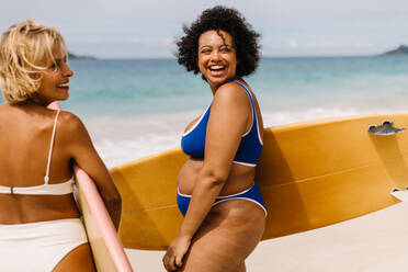 Zwei Frauen in Bikinis gehen an den Strand, um zu surfen, ihre Bretter in der Hand, bereit, die Wellen zu reiten und die Freiheit und Aufregung des Ozeans zu genießen. Surferinnen genießen das Strandleben. - JLPSF30655