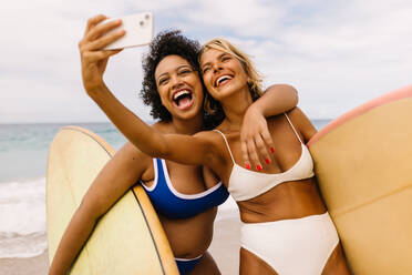 Best friends on a surfing vacation capture the moment with a joyful selfie on the beach, their smiles reflecting the exciting memories they create. Happy female surfers having fun together on shore. - JLPSF30659