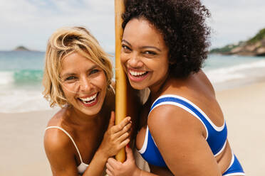 Two young women enjoy beach activities together, smiling happily on their summer surfing vacation. With a surfboard in hand, they embrace the thrill of surfing, creating lasting vacation memories. - JLPSF30664