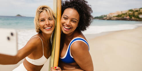 Two women in bikinis, one holding a surfboard, take a beach selfie with a camera phone. Happy and authentic, they celebrate their friendship on a fun vacation trip. Enjoying the ocean and surf, they create lasting memories. - JLPSF30665