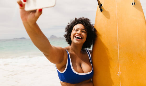 Eine fröhliche Plus-Size-Frau macht ein Selfie mit ihrem Surfbrett, während sie einen Solo-Sommerurlaub voller Strandaktivitäten und Erkundungen genießt - JLPSF30671