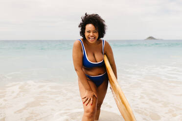 Junge Surferin, die sich am Strand vergnügt, ihr Surfbrett in der Hand hält und einen farbenfrohen Bikini trägt. Kurvige Frau, die in die Kamera lächelt, genießt einen aktiven Lebensstil und sommerlichen Solospaß. - JLPSF30673