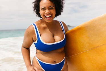 Körperbewusste Frau genießt einen sonnigen Tag am Strand, lächelt in die Kamera und trägt ihr Surfbrett. Weibliche Surferin erkundet den Strand mit Begeisterung und genießt das beste Sommererlebnis in ihrem Bikini. - JLPSF30675