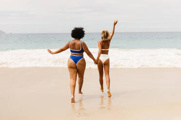 Frauen in Bikinis, die am Strand auf die Wellen zulaufen und den Geist des Sommerurlaubs zelebrieren. Zwei junge Freundinnen, die sich gemeinsam in einem aufregenden Strandurlaub vergnügen. - JLPSF30682