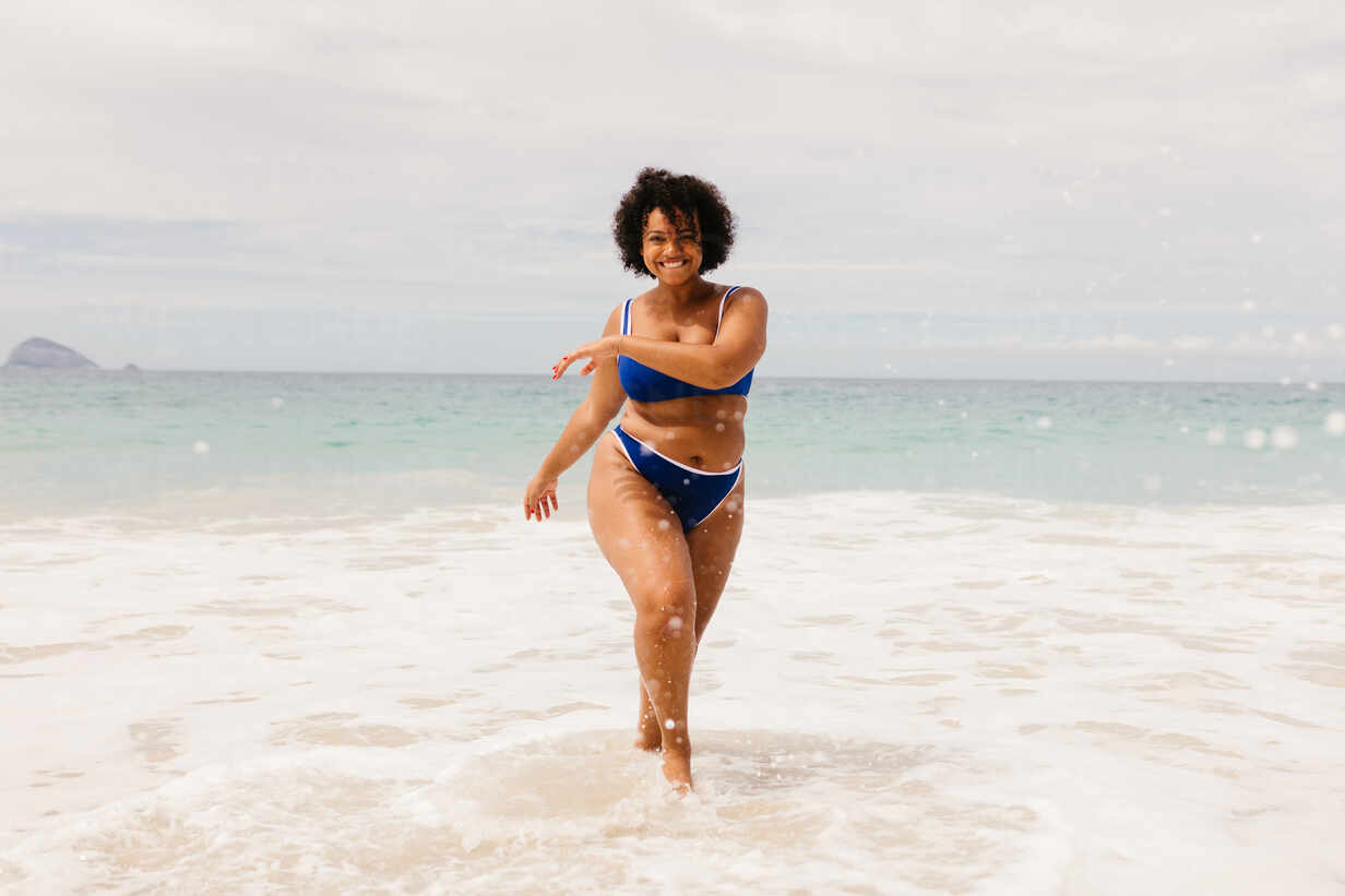 Happy young woman celebrating the spirit of summer on the beach, dancing  joyfully in her bikini. Body confident, plus size woman having fun on a  solo beach holiday. stock photo