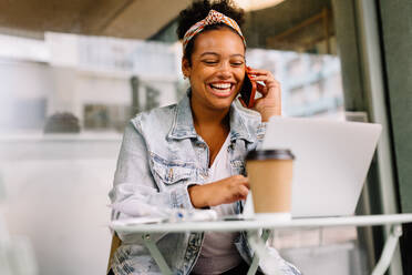 Happy businesswoman works remotely in a cafe, using her phone and laptop to communicate and stay productive. Her happiness reflects the freedom and fulfillment of freelancing. - JLPSF30695