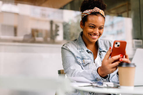 Fröhliche junge Frau, die in einem Café sitzt und mit einem echten Lächeln ihr Handy benutzt. Sie verkörpert den fröhlichen und authentischen Lebensstil einer Studentin oder eines Hochschulabsolventen. - JLPSF30697