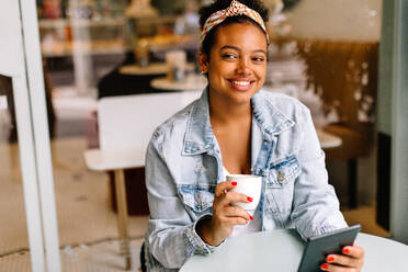 Junge Frau genießt ein Solo-Date in einem Café, hält einen heißen Kaffee in der Hand und benutzt ihr Tablet. Glückliche junge Frau, die sich in einem ruhigen Café entspannt und in den sozialen Medien surft. - JLPSF30701