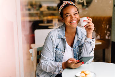 Fröhliche junge Frau genießt eine Tasse Kaffee in einem Café und surft mit einem Tablet in den sozialen Medien. Glückliche junge Frau, die ein authentisches Einzeldate genießt und einen entspannten und positiven Ausdruck zeigt. - JLPSF30704