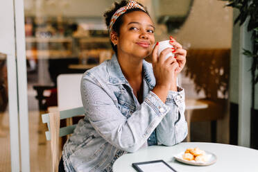 Eine junge Frau genießt einen Moment der Entspannung in einem Café, mit einem heißen Getränk in der Hand. Sie blickt gedankenverloren und mit nachdenklichem Gesichtsausdruck in die Ferne. Dieses echte und authentische Bild fängt die Essenz einer Verabredung zum Kaffee ein. - JLPSF30705