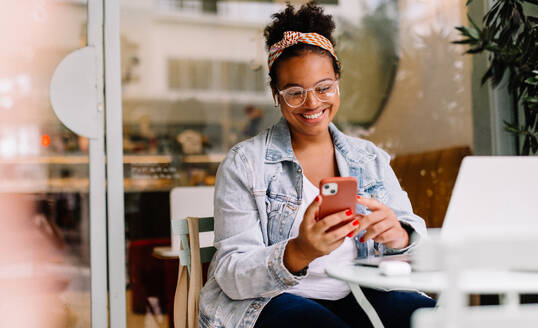 Happy woman works remotely in a café, using her smartphone for communication and social media. Her authentic smile reflects her happiness as a freelancer in the digital age. - JLPSF30707