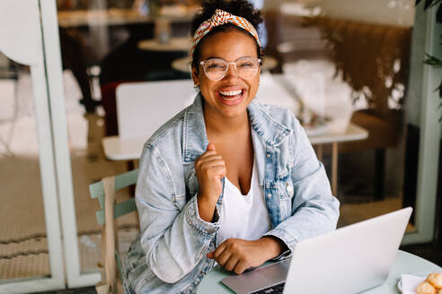 Young woman sits in a café, working on her laptop. She is a freelancer and content writer, enjoying the freedom of remote work. With a smile, she embodies the happiness of a successful entrepreneur. - JLPSF30715