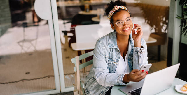 Cheerful young woman works happily in a coffee shop. She uses a laptop and mobile phone, showcasing the benefits of remote work. Her smile and focus depicts a confident entrepreneur in the digital age. - JLPSF30716